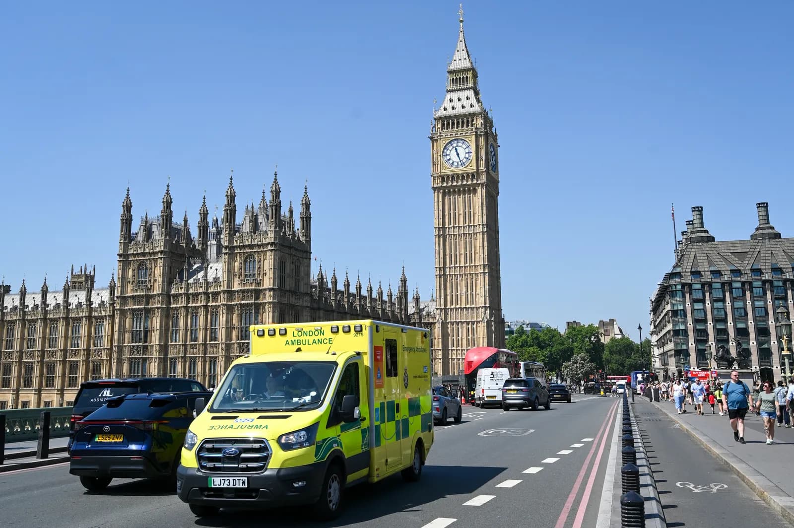 A bright yellow London Ambulance drives on a bridge with Big Ben and the Houses of Parliament in the background under a clear blue sky.